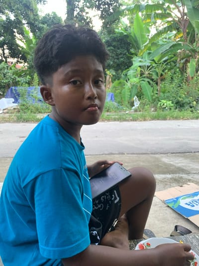The image displays a young male individual seated outdoors on a light grey concrete surface in Wedarijaksa, Indonesia. The individual, appearing to be a child or pre-teen, has a dark complexion and short, dark, curly hair with faded sides. He is wearing a plain, short-sleeved blue t-shirt and black shorts with white patterns. He looks directly at the camera with a neutral expression, his lips slightly pursed. His left hand is holding a black smartphone, resting on his left thigh, with the screen visible and displaying "16:45:25". On the ground to his right, a white plate with red and yellow floral patterns contains residual food. Adjacent to the plate, a partially visible blue and white cardboard box with the text "AGUA" and a pair of dark flip-flops are also present. The background features a narrow strip of grassy ground, followed by a grey paved road. Beyond the road, dense, lush tropical vegetation, including several large-leafed plants resembling banana trees and other green foliage, forms the backdrop. A blue tarp is partially visible amidst the foliage in the left background. The scene is illuminated by natural daylight.