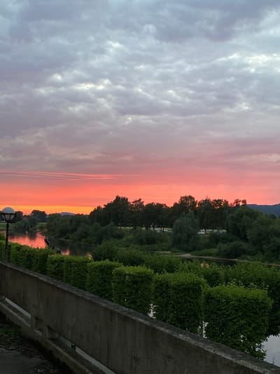 The image shows a sunset over the Weser River in Rinteln, Germany. The sky is filled with dramatic clouds, ranging from pale gray at the top to vibrant hues of pink and orange near the horizon. A calm river reflects the colors of the sky. Lush green trees and bushes line the riverbanks. In the foreground, a concrete wall and some manicured hedges create a foreground element. A streetlamp with a spherical shade is visible on the left side of the frame. The overall scene evokes a peaceful evening atmosphere.