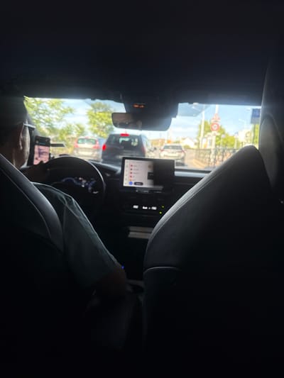 This is a first-person perspective from the back seat of a car in Nanterre, France. The driver, a person wearing a light blue shirt, is visible from the back, with their head and shoulder turned slightly to the left, holding the steering wheel. A smartphone mount with a phone is attached to the left side of the steering wheel. The car's dashboard is visible, with a digital display and a touchscreen infotainment system showing a list of icons and text. Outside the car, through the windshield, traffic is visible, including a silver car and a dark-colored SUV. Street signs, trees, and buildings are also visible in the background, suggesting an urban environment. The sky is blue with some clouds, indicating daytime. The interior of the car is dark, with the focus on the driver and the dashboard.