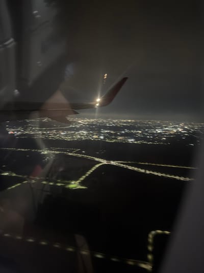 This is a photograph taken from an airplane window at night, looking down at a sprawling city illuminated by countless lights. The lights are blurred due to the movement of the aircraft, creating streaks of white and yellow against the dark landscape. A portion of the airplane's wing is visible in the upper left quadrant of the image. Roads, appearing as bright lines, crisscross the urban area. The city extends to the horizon, where it meets a dark, cloudy sky. The image does not depict Vellanki Agros, India, or any specific identifiable location within it.