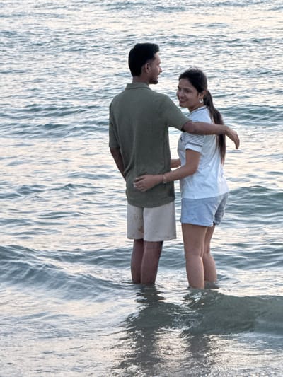 At Cenang Beach, Malaysia, two individuals are depicted standing in the shallow sea water, which reaches approximately mid-calf. The male, positioned on the left, is wearing a dark olive green short-sleeved collared shirt and light beige shorts. His left arm is extended around the female's waist, and he is looking towards her. The female, positioned on the right, is wearing a white short-sleeved t-shirt with thin horizontal stripes on the sleeves and light blue shorts. Her right arm is extended around the male's lower back/waist. She possesses dark hair, tied back in a ponytail, and is wearing white hoop earrings. The female is observed looking directly towards the viewer with a smile. The surrounding water exhibits gentle ripples, and the overall lighting is soft, suggesting either early morning or late afternoon. The background consists solely of open water, with no distant land visible.