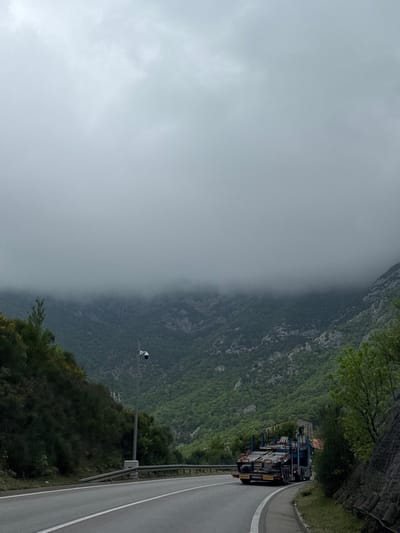 An outdoor, rotated image captures a mountainous landscape partially obscured by heavy fog or clouds. The scene is dominated by a road with a white solid line on the shoulder and a broken white line separating lanes, curving gently to the right in the foreground. A metal guardrail runs alongside the road.
On the right side of the road, a large semi-trailer truck is visible, laden with what appears to be construction materials, possibly scaffolding or pre-fabricated components, stacked high and secured to a flatbed trailer. The truck is parked or moving slowly on the side of the road.
To the left of the road, a tall grey pole stands with a white camera or sensor device mounted on top, facing towards the road or the landscape beyond. Lush green vegetation, including trees and bushes, covers the steep slopes of the mountains that rise directly from the roadside. Further up the mountain on the right, dark grey protective netting is visible against the rock face, suggesting measures against rockslides.
The mid-ground and background are enveloped in a thick layer of grey fog or low clouds, which obscures the upper parts of the mountains and creates a hazy, muted atmosphere. The mountains themselves are rugged, with visible rock formations interspersed with dense green foliage. The time of day appears to be daytime, but the overcast conditions make the light diffused. No people are explicitly visible, and there is no discernible text. The scene depicts a journey through a rugged, perhaps challenging, mountain pass in Đurmani, Montenegro.