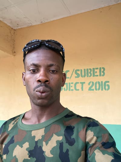 A dark-skinned man in a camouflage shirt poses, wearing sunglasses on his head. He has a trimmed goatee and mustache. The background features a wall painted yellow with green text that reads "C/SUBEB PROJECT 2016." The wall also has a green stripe towards the upper right corner. The man appears to be positioned close to the camera, emphasizing his face and upper body.