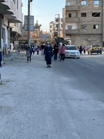 This image depicts a daytime street scene in Mu'adammiyat ash-Sham, Syria.
In the foreground, the ground on the left consists of uneven, dusty terrain with scattered debris and small piles of dirt. This transitions into a paved asphalt or concrete road surface on the right, exhibiting minor cracks and dust.
Approximately 15-20 individuals are present. In the midground, slightly left of center, a female adult wearing a dark blue abaya and a light-colored headscarf walks towards the camera, holding a dark bag. To her right, a group of three female adults walk together; one wears black, and two wear pink tops. Further down the street, multiple children, most carrying backpacks, are visible walking on both sides of the road, suggesting they are school-aged. A young boy with a light-colored backpack is walking away from the camera on the unpaved left side. Other adults are present in the background.
Vehicles include a white sedan positioned on the right side of the road, behind the group of women, and a yellow taxi cab parked further down the street on the right.
Multi-story buildings constructed from concrete blocks and masonry line both sides of the street. Structures on the left exhibit signs of partial construction or disrepair, with exposed rebar and unfinished sections. Buildings on the right display worn facades, darkened window openings, and one external air conditioning unit. A prominent utility pole with attached signage stands on the left side of the paved road. The sky above is clear.
