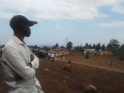 A man in a white shirt and a black cap stands in the foreground with arms crossed, looking off to the left. Behind him is a field of dark soil. In the background, there is a group of people, cars, and yellow buses. There are also buildings, trees, and an electric power line visible. The sky is light blue with white clouds.