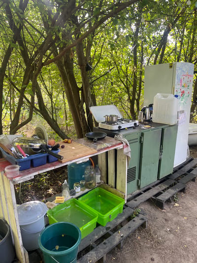 An outdoor kitchen installation is observed within a densely wooded environment in Sint-Pieters-Leeuw, Belgium. The scene is illuminated by natural light filtering through a dense canopy of green foliage. The setup includes a makeshift counter constructed from light-colored wooden planks, supporting a blue plastic dish rack filled with assorted kitchenware, a wooden cutting board, and several pieces of fruit. Below this counter, a blue gas cylinder, various bottles, and glass jars are visible. On the ground directly beneath, two bright green plastic tubs filled with water, a yellow bar of soap, and a brush are present. Adjacent to this section, a darker counter surface holds a white portable gas stove with a single burner, on which a silver pot rests. A black frying pan, a silver Moka pot, another small dark container, and a large clear plastic jerrycan with a blue cap are also on this surface. To the far right stands a light-colored upright refrigerator, adorned with colorful drawings and magnets. The ground beneath the entire assembly consists of unpaved earth, partially covered by dark wooden pallets serving as a base. No individuals are present within the frame.