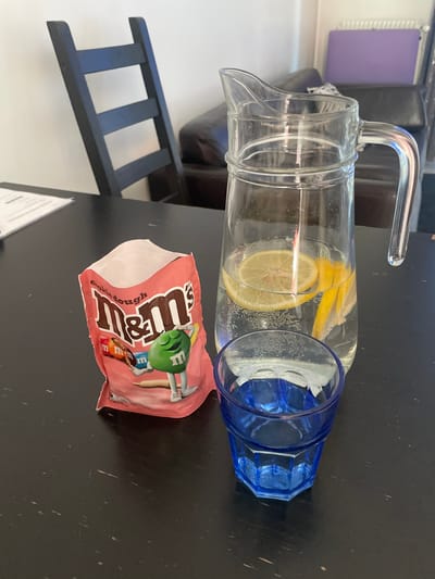 On a dark, wooden table, a bag of M&M's cookie dough candies is positioned next to a glass pitcher filled with water and lemon slices. A blue glass sits in front of the pitcher. In the background, a dark wooden chair with horizontal slats is visible, along with a plush brown sofa and a purple bulletin board. The setting appears to be an indoor domestic environment, possibly a living room or dining area, in Vénissieux, France.