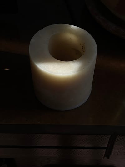 The image features a solitary, cylindrical object, likely a solid tube or a hollowed-out block, predominantly off-white or cream in color. The object is oriented horizontally, with its circular opening facing towards the left of the frame. It appears to be made of a dense, opaque material, possibly wax, plastic, or ceramic, and has a slightly textured surface with minor imperfections or dust specks visible, particularly on the inner rim and walls of the hollow center.
A strong, directional light source illuminates the object from the left, casting a bright highlight on the left-facing edge and the interior surface of the cylinder. This lighting creates a dramatic contrast, with the left side of the object brightly lit and gradually fading into shadow towards the right side. The right end of the cylinder is largely obscured by shadow.
The background is very dark and mostly indistinct, emphasizing the illuminated object. To the far right, there are glimpses of dark, vertical elements, possibly parts of a wooden structure or furniture, exhibiting some subtle texture. The surface beneath or behind the object is also dark, blending into the background. There is no visible text or discernable activity. The overall impression is one of a focused, artistic study of form and light on a simple, everyday object within a dimly lit environment.