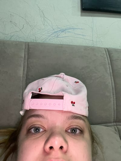 A close-up, overhead, indoor shot captures a person's face and the top of a pink baseball cap with cherry patterns. The cap is tilted forward, obscuring the forehead and the top of the head. The person's eyes are open and looking upward. The background consists of a textured, light-colored wall with visible scuff marks and a portion of a gray upholstered surface, likely a sofa or bed. The image does not contain any contextual information to determine its location within the city of Gorodets, Russia.