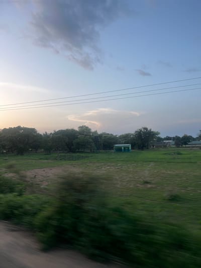 This image depicts a rural or semi-rural landscape in Ribina East, Nigeria, captured during what appears to be late afternoon or early evening. The scene is characterized by a wide sky transitioning from a pale, light yellow-orange on the left horizon to a clearer, light blue overhead and a deeper blue with darker grey clouds on the right. Three parallel power lines stretch horizontally across the upper middle section of the frame.

The foreground is blurred with streaks of green and brown, consistent with motion blur, suggesting the photograph was taken from a moving vehicle. This blurred area transitions into a broad expanse of green field in the midground, with a patch of lighter, dry earth visible on the left side.

In the background, a dense line of various green trees forms a natural barrier. Interspersed with and behind these trees are several structures. A distinct, small, rectangular building with a flat roof, painted in two shades of green (lighter on top, darker on the bottom), is visible in the middle-right. Further to the right and behind the trees, additional buildings, possibly residential or institutional, are partially discernible. A dark water tank, supported by a metal structure, is partially visible above the treeline on the right. No people or animals are visible, and no specific actions or interactions are depicted within the frame.