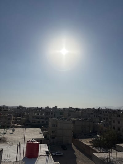 The image displays an elevated, wide shot of a dense urban environment in Mu'adammiyat ash-Sham, Syria, under a clear, bright sky. A prominent sunburst, indicative of direct sunlight, is visible in the upper-center of the frame.
The cityscape consists primarily of multi-story buildings constructed from concrete and masonry, appearing in shades of gray, beige, and light brown. Many structures exhibit visible signs of damage, decay, or incomplete construction, with exposed rebar protruding from rooftops and walls, and missing window sections. Buildings are densely packed, forming narrow gaps and unpaved pathways at ground level.
In the lower-left foreground, a concrete rooftop features a red, cylindrical water tank. To its left, another concrete rooftop has a white satellite dish and several vertical rebar elements. Below these rooftops, a dirt path or unpaved road shows two parked vehicles: a light-colored sedan (likely white) in the foreground and a darker vehicle behind it. Sparse, green vegetation is visible between some buildings towards the right. The distant horizon reveals faint, hazy hills or mountains. No individuals are depicted, and no specific actions or interactions are discernible.