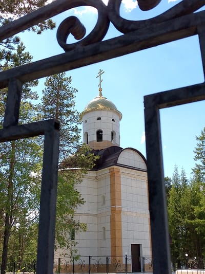 The image shows the Church of the Holy Trinity in Neryungri, Russia. The church is a white, multi-story building with a golden dome and a cross on top. It is surrounded by trees and a black metal fence. The sky is blue with some clouds. The church appears to be a modern structure, likely built in the late 20th or early 21st century, judging by its design and construction materials. The fence in the foreground is made of wrought iron with decorative elements. The overall scene suggests a peaceful and serene setting.