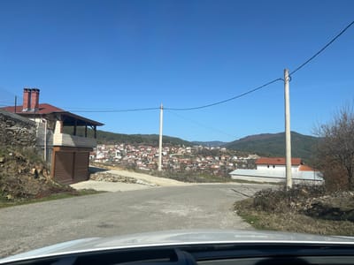 The image depicts a rural or semi-rural street scene in Oreshe, Bulgaria, on a clear, sunny day. The perspective appears to be from inside a vehicle, with the upper portion of a dashboard visible at the bottom. A paved road curves slightly uphill and to the right, leading into the mid-ground.

On the left, a two-story building stands with a red-tiled roof featuring two brick chimneys. The upper level includes a covered wooden balcony, while the lower level features two brown, roll-down security doors or garage doors. Adjacent to this building on the left is a dry-stacked stone retaining wall and an unkempt grassy bank.

In the mid-ground, a tall, light-colored utility pole stands to the right of the road, supporting a black electrical wire that stretches horizontally across the frame. Beyond this pole, on the right side of the road, is another building with a white facade and a red-tiled roof, partially obscured by bare, dormant vegetation.

The background reveals a dense village or townscape situated in a valley, with numerous houses displaying red-tiled roofs. A white minaret is discernible among these structures, indicating the presence of a mosque. The village is surrounded by hills and mountains, featuring a mix of coniferous and bare deciduous trees under a vibrant, cloudless blue sky. No individuals, overt actions, or direct interactions are visible.