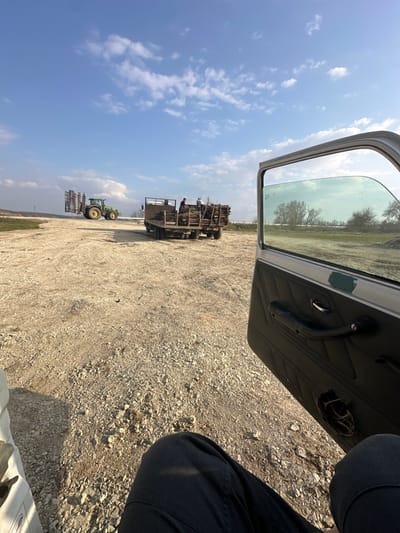 The scene is set in an outdoor, rural or semi-rural environment in Dolna Studena, Bulgaria, observed from the interior of a parked vehicle with its passenger door open. In the immediate foreground, the lower legs of an individual wearing dark trousers are visible, positioned towards the lower right. The open vehicle door, located on the right side, displays a dark-colored interior panel with a textured surface, a chrome door handle, and a manual window crank. The window glass is partially lowered, reflecting green light. The midground is dominated by an expanse of light-colored, unpaved ground composed of dirt and gravel. Centrally positioned in the midground is a flatbed truck, laden with a substantial quantity of cut wood or tree branches. Two individuals are present on the truck's bed; one is standing near the center, facing left, and the other is further towards the rear. To the left of the truck, a green tractor, equipped with a large, multi-tine agricultural implement attached to its rear, is parked. The background features a flat horizon line with distant, sparse vegetation and trees under a clear blue sky with scattered white cumulus clouds.