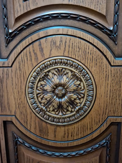 The image displays a close-up view of decorative wooden paneling. The central focus is an ornate circular carving with a floral motif, rendered in a darker, possibly metallic, finish against the natural wood grain. The wood itself has a visible grain pattern and a warm, brown tone, likely oak. Surrounding the central motif are concentric carved borders and architectural elements, suggesting it's part of a larger piece of furniture or interior design, such as a cabinet door or a wall panel. The lighting highlights the three-dimensional aspects of the carvings, creating shadows and emphasizing the depth of the design. There are no people, actions, or environmental context beyond the object itself. The location in Nalchik, Russia, is not discernible from the visual information presented in the image.