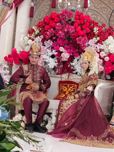 In Meuliek, Indonesia, a wedding ceremony takes place indoors, possibly in a hall decorated with numerous vibrant floral arrangements. At the center of the scene, a newlywed couple is seated on an ornate couch. The groom, on the left, is dressed in a maroon traditional outfit adorned with intricate gold embroidery and a golden headdress. He is holding a small yellow object in his hands. The bride, on the right, wears a similar maroon attire, also heavily embellished with gold. Her headdress is elaborate and golden, complemented by a veil that drapes over her shoulders. Both individuals appear to be observing the proceedings. The background is dominated by a lush backdrop of red, pink, and white flowers, with a decorative architectural element and a chandelier visible above. The overall ambiance suggests a festive and formal occasion.