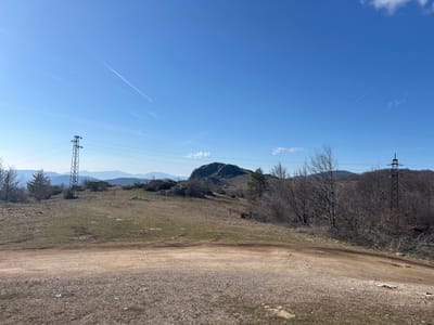 The scene depicts an open, elevated landscape in Ichera, Bulgaria, under a clear blue sky with two prominent white aircraft contrails. The foreground consists of a light brown, unpaved dirt path or road, sparsely covered with gravel and small rocks, curving from the lower left towards the lower right. The terrain immediately beyond the path is characterized by gently sloping hills covered in dry, sparse greenish-brown grass and scattered rock formations.
In the left midground, a tall, metal lattice-work electricity transmission tower is positioned, supporting multiple parallel power lines that extend horizontally across the frame. A second, smaller transmission tower is partially visible on the far right, amidst bare-branched deciduous trees. A distinctive, rounded rocky mound or hill dominates the central midground. Sparse vegetation, including scattered bare deciduous trees and a few evergreen trees, is present on the hillsides. Distant mountain ranges appear as pale blue silhouettes against the horizon. No human or animal subjects are visible. A small, reddish-orange marker with a white top is barely discernible on the grassy slope to the right of the central rocky mound. The lighting suggests a bright, sunny day.