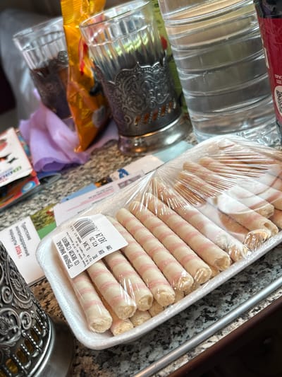 The image depicts a close-up view of a table setting with several objects. In the foreground, a plastic-wrapped package of wafer rolls with pink stripes is prominently displayed. To the left, a decorative silver goblet or holder is visible. Behind the wafer rolls, a clear plastic water bottle with ribbed detailing and a red label is present. In the background, two glass tumblers, one appearing to contain a dark liquid, sit next to a bright yellow package. A crumpled purple cloth lies between the glasses and the tumblers. On the table surface, several papers or leaflets are scattered, including one with visible Cyrillic text and a date, "2026 г." The overall setting appears to be indoors, possibly a home or a cafe. The specific location in the city of Kotelnich, Russia, is not directly indicated by any visual cues in the image.