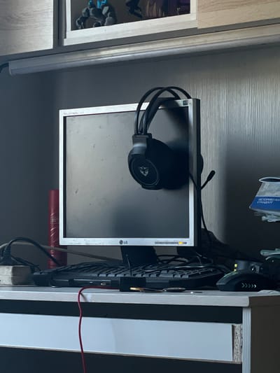 The image depicts a desk with a computer monitor, keyboard, mouse, and headphones. The monitor is a flat-screen display with the LG logo visible at the bottom. Headphones are hanging on the side of the monitor. A keyboard is positioned in front of the monitor, and a computer mouse is to the right of the keyboard. A red cable is visible on the desk. In the background, shelves hold figurines and a blue booklet. The overall environment appears to be a home office or a personal workspace. The location is specified as Новая Тура, Russia.