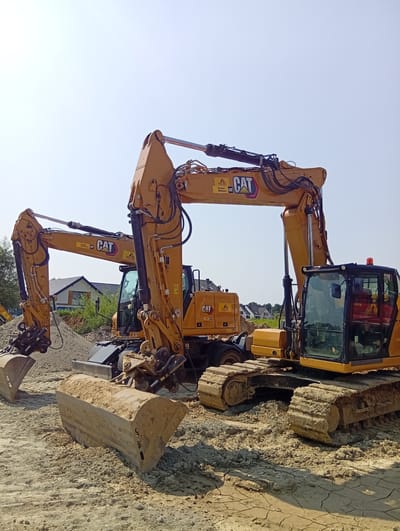 The image shows two yellow Caterpillar excavators on a construction site, likely in Długołęka, Poland, given the context. The excavators are positioned in a dirt area with piles of sand and loose soil. The excavator in the foreground has tracks that are heavily covered in mud, and its bucket is partially buried in the dirt. The second excavator, located behind and to the left, also appears to be on tracks and has its arm extended. Both machines display the CAT logo. In the background, residential houses with tiled roofs and trees are visible under a clear, bright sky. The scene suggests ongoing construction or landscaping work. No people are visible in the image.