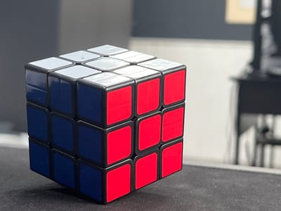 A Rubik's cube sits on a dark, textured surface, possibly a desk or table. The cube is partially solved, with the top face showing white stickers, the left face displaying blue stickers, and the front face featuring red stickers. The background is out of focus, showing a gray wall and lighter colored objects further back, including what appears to be furniture. The lighting suggests an indoor environment. There is no indication of Kalininets, Russia, in the image.