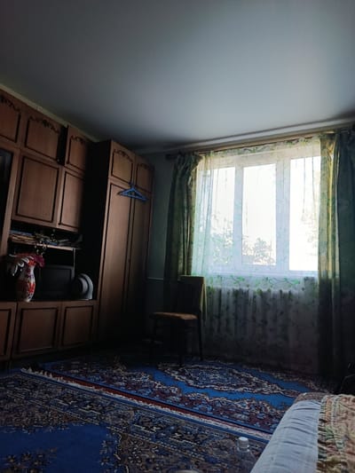 The image depicts an indoor scene, likely a bedroom or living room, in what appears to be a residence in Rohizna, Ukraine. The room is furnished with a large wooden wardrobe on the left, which has multiple doors and shelves. On one shelf, a vase with flowers and a small television are visible. A blue hanger is placed on one of the wardrobe doors.
A window with white panes is central to the image, covered by sheer floral-patterned curtains in green and white. Outside the window, trees and sunlight are visible, suggesting daytime. A radiator is positioned below the window. To the right of the window, a green curtain is partially drawn.
In front of the window and to the right, a chair with a wooden frame and a dark seat is placed. The floor is covered by two Persian-style rugs with intricate blue, red, and brown patterns. Part of a bed with white and patterned bedding is visible in the lower right corner of the image. The ceiling is smooth and white. The lighting suggests natural light streaming through the window, casting shadows within the room. The overall atmosphere is that of a lived-in, traditional interior.