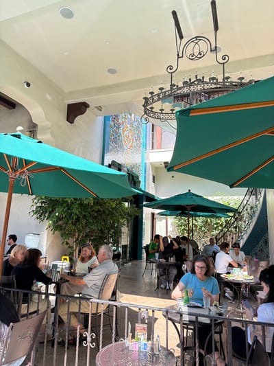 The image shows an outdoor dining area at The Raymond 1906 restaurant, located at 1250 South Fair Oaks Avenue, Pasadena, United States of America. Several patrons are seated at tables, enjoying meals and drinks. Large teal umbrellas provide shade. A decorative chandelier hangs from the ceiling. The restaurant features an ornate tiled wall.
