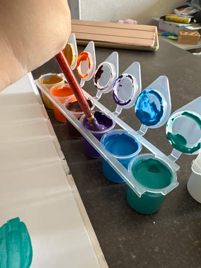 A hand holding a paintbrush is shown dipping the brush into a small container of purple paint. The paint pots are arranged in a white plastic holder, and there are various colors of paint visible, including orange, yellow, brown, purple, blue, and green. A white sheet of paper is positioned to the left of the paint pots, with some green paint already applied to it. The scene appears to be set on a dark, speckled countertop. In the background, there are some objects on shelves, including what looks like a wooden block and some boxes. The overall impression is of someone engaged in painting or a related art activity. The image does not provide any information about the location being the city of Верхнерусское, Russia.