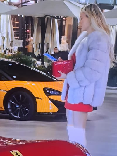 A blonde woman wearing a fluffy white coat over a red dress stands next to a bright orange McLaren sports car. She holds a red clutch bag. Behind her, several other people are visible, and the background appears to be an outdoor seating area with umbrellas and foliage. A portion of a red Ferrari is visible in the foreground. The scene is likely in an urban setting, though specific details of Cēsis, Latvia, are not discernable from the image.