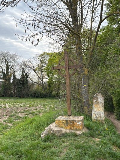 The image shows a Celtic cross, possibly made of wood, standing on a stone base in Auvers-sur-Oise, France. The cross and the stone base are covered in moss and lichen. The cross is located in a grassy area with trees in the background. The sky is overcast. Another stone structure is visible next to the cross, likely part of a cemetery or historical site. The surrounding vegetation suggests a rural or semi-rural environment.