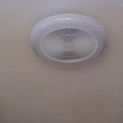 A white, circular fan is mounted on a beige ceiling. The fan has a ridged grille with radiating lines. The ceiling appears to be textured. The image is a close-up shot of the fan and ceiling, with the fan taking up the upper portion of the frame. The lower portion of the image is entirely the beige ceiling.