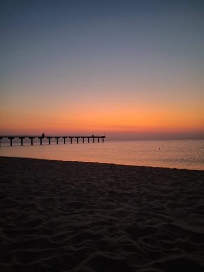 The image depicts a tranquil beach scene at dawn or dusk in Shkorpilovtsi, Bulgaria. In the foreground, the dark, rippled sand of the beach fills the lower portion of the frame, creating a stark contrast with the brighter elements in the distance. Extending out over the calm, reflective water is a long, silhouetted pier, supported by numerous pilings. The pier stretches horizontally across the middle ground, disappearing into the distance. The sky above transitions from a deep, dark blue at the top to a vibrant gradient of orange and pink near the horizon, indicative of sunrise or sunset. The water is a muted orange-pink, mirroring the sky. There is a small, dark spherical object visible in the water to the right of the pier. A small structure, possibly a lifeguard station, is visible on the pier. No people are discernible in the image. The overall atmosphere is serene and still.