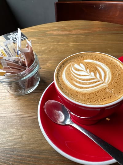 The image depicts an indoor scene within a cafe environment in Chester, United Kingdom. In the foreground, a red ceramic coffee cup with a matching saucer holds a latte featuring a distinct white fern or leaf pattern in its foam. A silver spoon rests on the right side of the saucer. To the left of the cup, a clear glass jar contains an assortment of individual sugar and sweetener packets, including varieties labeled 'Sucralose,' 'Sweetener,' and 'Sugarcane.' All items are positioned on a light brown wooden table exhibiting grain patterns and minor wear. In the upper left corner, a partially visible, inverted black card with a QR code and the word 'SCAN' is present. The background consists of a dark grey wall, with a segment of a reddish-brown wooden chair visible on the far right. No individuals or active interactions are depicted.
