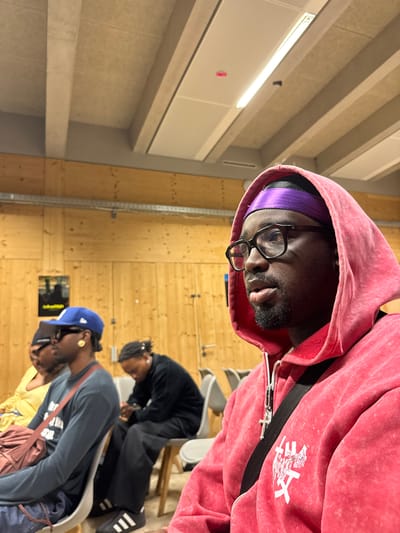 A medium shot depicts a Black male wearing a pink hoodie, glasses, and a purple head covering. He is positioned in the foreground of the image. Behind him, in the middle ground, are two other individuals seated on chairs. One is wearing a blue baseball cap and a grey long-sleeve shirt, while the other is wearing all black. The background reveals a room with light-colored walls and ceiling fixtures. Based on additional information, the location of the scene is within the city of Saint-Ouen-sur-Seine, France. The individuals are interacting in an indoor setting, possibly a classroom or studio.