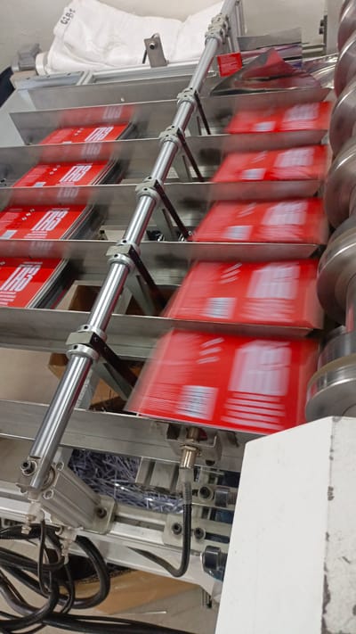 The image shows a close-up, slightly angled shot of a conveyor belt system in an industrial setting. Stacks of red-covered booklets or pamphlets are being processed on tiered metal shelves. The items appear to be moving from left to right. The equipment is primarily made of metal, with visible chrome-plated posts, brackets, and rollers. There are also black cables and electrical components present. In the background, a large white bag with some text and markings is visible, along with what appears to be shredded paper or material. The overall scene suggests an active production or packaging line. The location is not specified in the image or the provided crops, therefore the city of Meget, Russia, cannot be determined.