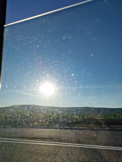 The image captures an outdoor scene from the perspective of looking out a vehicle's side window, rotated 90 degrees clockwise from its likely original landscape orientation. When viewed correctly, a bright blue sky dominates the upper portion, featuring a strong, brilliant sun causing significant lens flare and numerous bokeh effects, which appear to be dust particles or water droplets on the windowpane.
Below the sky, a rural landscape unfolds with a gentle horizon line revealing rolling hills or elevated terrain covered in green vegetation, indicative of fields or natural growth. On the far right, a dark asphalt road with two distinct parallel white lines is visible in the foreground, suggesting a highway or main road. The imperfections of the glass surface, including reflections and specks, are noticeable throughout the image. The scene depicts a clear, sunny day, likely during the mid-morning or afternoon, indicating travel through a rural area. No visible text is present. The setting is Tatarka, Russia.