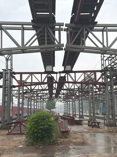 The image depicts an outdoor scene at a train station under construction in Ballabgarh, India. The foreground features a muddy, wet ground with scattered debris and a small green bush. A wooden bench with a concrete base is visible to the right of the bush. Beyond this, a train with red carriages is stopped at the platform. The background is dominated by the skeletal framework of what appears to be an elevated walkway or station roof, constructed from grey and red metal beams. The sky is overcast and a light haze is present. No people are clearly identifiable in the scene, though indistinct figures are visible further down the platform. The overall impression is one of ongoing construction at a functional train station.