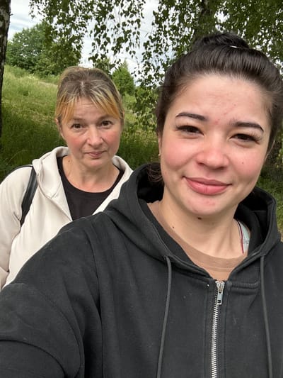 In the city of Gavrilov Posad, Russia, two women are captured in a selfie outdoors. The woman in the foreground, closer to the camera, has her dark hair pulled up in a bun and is wearing a black zip-up hoodie over a tan t-shirt. Her eyes are dark and she has a slight smile. The woman in the background, with blonde hair pulled back, is wearing a white zip-up jacket over a black t-shirt and has a neutral expression. Both women are positioned against a backdrop of lush green foliage, with trees and grass visible. The sky above appears overcast.