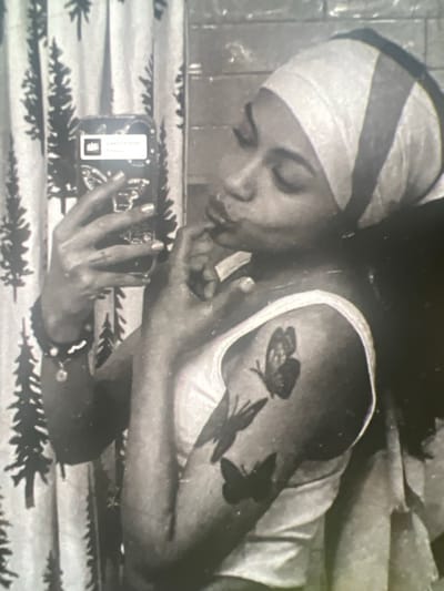 A black and white, medium-shot, selfie-style photograph shows a young Black woman in profile, looking into her phone as she takes a picture. She is wearing a light-colored tank top and a headscarf with dark stripes. She has a tattoo of three butterflies on her left arm. Her right hand is raised to her face, with her fingers touching her lips. In her left hand, she holds a phone with a butterfly design on the case. The background is a shower curtain with a pattern of trees. The image appears to have been taken in a bathroom. The exact location in the city of Boone, United States of America, is not discernible from the image.