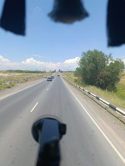 The image shows a view from inside a vehicle traveling on a highway. The road is asphalt with clear white lane markings. Several cars are visible on the road, traveling in the same direction. The landscape on either side of the road is dry and grassy with scattered rocks and some vegetation. A line of trees is visible in the distance to the right, and a larger tree is prominent in the middle right of the image. The sky is blue with scattered white clouds. In the foreground, a black object, likely part of the vehicle's interior or a mount, is out of focus. The scene appears to be in a rural or semi-rural area, consistent with the location of Dzoraghbyur, Armenia, which is known for its natural landscapes and proximity to highways.