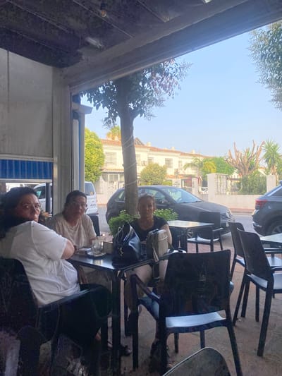 The image depicts an outdoor seating area at a cafe or restaurant in San Pedro De Alcántara, Spain. Three women are seated at a table. The woman on the left, wearing a white t-shirt and dark pants, is looking towards the camera. The woman in the center, wearing a light-colored patterned t-shirt and glasses, is also looking forward. The woman on the right, wearing a dark t-shirt, is looking to her left. A table is set with drinks, a pack of cigarettes, and bags. In the background, there are parked cars, including a black Volkswagen Golf and a dark SUV, and residential buildings with white facades. Trees and foliage are visible, and the sky is clear blue. The scene appears to be captured during daylight hours.