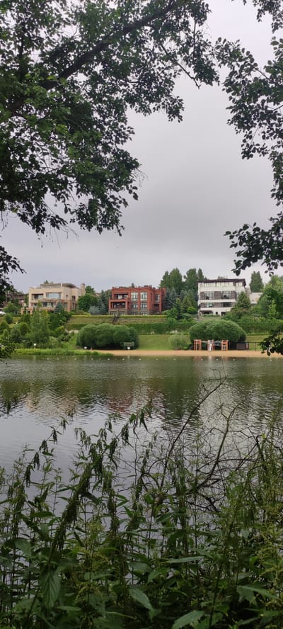 This media file captures a serene lakefront scene in Angelovo, Russia. In the foreground, lush green foliage, including nettles, partially obscures the view of the calm water. The lake's surface reflects the overcast sky and the structures on the opposite bank.
Across the water, a sandy beach area is visible, featuring a playground with a slide and climbing structure, a volleyball net, and a few small buoys marking the swimming area. Beyond the beach, a manicured grassy slope rises, dotted with various trees and shrubs.
Perched on the slope are three modern houses. The leftmost house is a light-colored, multi-story building with numerous windows and balconies. To its right stands a distinctive red brick house with a more industrial aesthetic, featuring large, recessed windows and multiple levels. The third house, on the far right, is a white, multi-story structure with a flat roof and many windows, appearing more contemporary.
The sky is uniformly gray, indicating an overcast day. The diffused light suggests it might be morning or afternoon. There are no people visible in the scene, contributing to the tranquil and uninhabited feel. The overall impression is one of peaceful residential living adjacent to a natural recreational area. No visible text is present in the image.