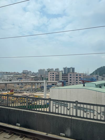 This image captures a cityscape with a focus on urban development and infrastructure. The scene is dominated by a dense collection of multi-story buildings, some appearing modern with numerous windows, while others show signs of ongoing construction. Several cranes are visible among the buildings, indicating active construction sites. The foreground features a metallic railing and a concrete structure, possibly part of an elevated walkway or bridge. Power lines run diagonally across the sky, which is overcast with scattered clouds, suggesting a potentially humid or hazy day. The perspective is from a slightly elevated position, looking out over the city. The overall impression is one of a rapidly developing urban area in Dongguan, China, with a blend of existing structures and new projects underway. There is no visible text in the image.