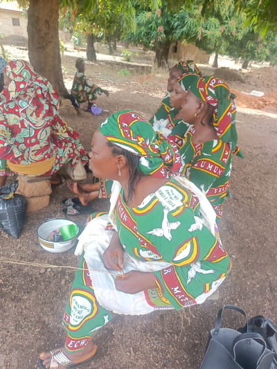A group of African women are gathered outdoors in Ninzo South, Nigeria. They wear matching green, white, and red patterned fabric dresses and headwraps. One woman in the foreground sits on a low seat, possibly a brick, and appears to be manipulating thin sticks or fibers in her hands. A metal bowl and a black plastic bag sit near her. A black handbag rests on the ground near her feet. Other women are seated behind her, and one is seated near a tree in the background. The ground is dirt and there are trees overhead. The women are likely attending a gathering or event, as indicated by their matching attire.