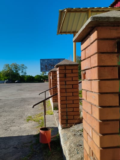 This outdoor photograph, taken in Velyka Bahachka, Ukraine, captures a sunny day scene. In the foreground, a series of sturdy brick columns with concrete caps define a boundary or entrance. A dark metal handrail extends from one of these columns, leading down towards an orange, cylindrical metal bucket with legs, resting on a patch of grass and dirt next to the paved ground. The ground transitions from a coarse, concrete-like material to a larger paved area, possibly an asphalt parking lot or road, which stretches into the mid-ground.

The sky is a vibrant, clear blue, indicating bright, sunny weather. Long, distinct shadows are cast across the paved surface, suggesting it is midday or early afternoon. In the background, beyond the paved area, a line of dense green trees and foliage is visible, with two dark vehicles partially obscured underneath them.

To the right, part of a building's roof, featuring yellow corrugated metal and a red-tiled peak, is visible. A rectangular, weathered blue sign or billboard, showing signs of peeling and faint, largely illegible white markings, stands in the mid-ground. A small white arrow symbol is visible on this sign. The brick columns themselves show some minor wear and what appears to be faint green graffiti on one side. No people are present in the image.