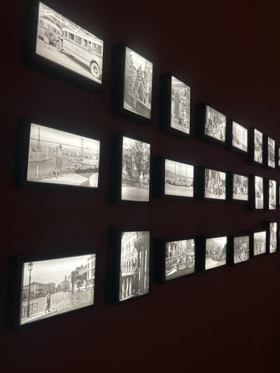 The image displays a gallery of backlit black and white photographs mounted on a dark red wall. The photographs are arranged in a staggered pattern, creating multiple rows. In the top row, the leftmost photograph shows a vintage bus with passengers. The subsequent photographs in this row depict architectural scenes and street views. The second row from the top features images of people and street scenes, including a man standing on a walkway with a suspension bridge in the background. Further to the right, a photograph shows palm trees and two individuals. The third row from the top contains images of boats docked at a harbor, a group of people in an outdoor setting, and architectural elements. The bottom row starts with a photograph of a man standing on a street next to a canal with buildings lining the waterway, reminiscent of Venice. This image is followed by photographs of architectural facades and street scenes. The overall impression is of a curated collection of historical images, likely documenting urban life and historical sites. The reference to Trakai, Lithuania is not visually supported by the provided image.