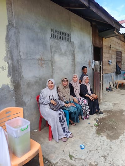 In Sangso, Indonesia, four women and one young male are gathered outside a building. The women are seated on red and blue plastic chairs in front of a gray, weathered concrete wall. Three of the women are wearing headscarves and modest dresses, with one woman holding a handbag. The young male stands behind them, looking towards the camera. To the left, an orange plastic chair holds a large clear container filled with a light green liquid. The building behind them appears to be constructed of wood and corrugated metal. The ground is a mixture of cracked concrete and dirt.