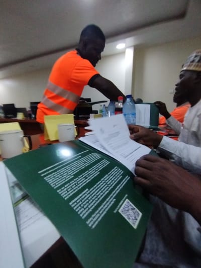 The image shows a meeting taking place at an office in Ashaka, Nigeria. Two men are seated at a table. One is wearing an orange shirt with a reflective vest. The other man, sitting to the right, is wearing a white shirt. The man in the orange shirt is standing up, leaning over the table as if addressing the other man who is seated with a green document in his hands. Multiple water bottles and a Coca-Cola bottle are visible on the table, along with a clipboard, a yellow mug, and a white mug. A few computer monitors are in the background. The image is taken from a low angle, with the focus on the green document.