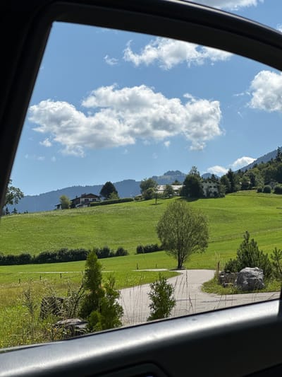 The image displays a view through a car window, looking out onto a sunlit, green countryside. The foreground features a portion of the car's interior, a dark grey door panel, obscuring the bottom of the scene. A paved path, curving from the right side of the image towards the center, is visible. To the left of the path, wild grasses and small yellow flowers grow, interspersed with rocks and a few small evergreen trees. Further into the scene, the path meanders through a vibrant green, rolling meadow. Several trees are scattered across the meadow, with a larger, leafy tree prominently positioned in the middle ground.

In the background, a cluster of traditional alpine-style houses with red roofs are nestled on a hillside. The hillside is covered in lush green grass, with some lighter patches that could be wildflowers or shorter grass. Behind the houses, a dense forest covers the slopes of a large mountain range, which stretches across the horizon. The sky above is a clear, bright blue, dotted with fluffy white cumulus clouds. The overall impression is a peaceful, rural landscape on a clear, sunny day, likely in a mountainous region. Based on the architectural style of the houses and the landscape, this scene is consistent with the region of Bischofswiesen, Germany, known for its picturesque Bavarian Alps scenery. No people are visible in the image.