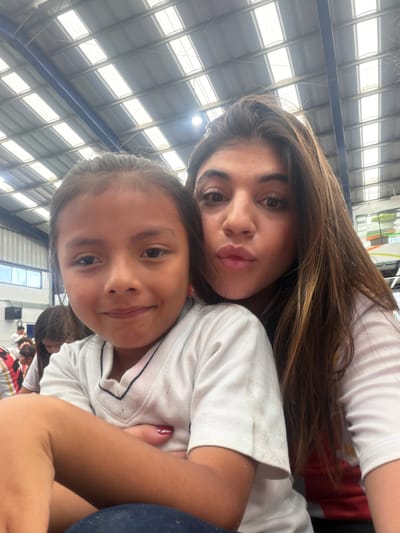A selfie shows two females, one younger than the other, indoors, in what looks like a gymnasium in Tlaxcalancingo, Mexico. The younger female is in the foreground; she has dark blonde hair tied into pigtails, fair skin, and is smiling. The older female, directly behind and above her, has long, light brown hair and is also smiling. Both appear to be wearing white shirts. Part of the gymnasium structure is visible in the background. The ceiling is a grid-like structure made of steel beams and translucent panels. Other people are visible further back in the gymnasium.